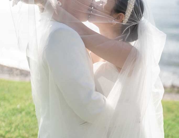 A bride and groom share a kiss, wrapped in a veil with Lake Como and the Alps in soft focus behind them.