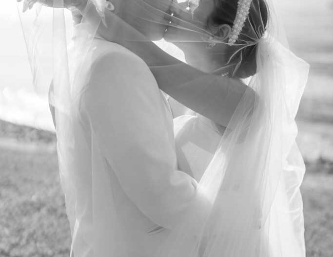 n a black and white photo, a bride and groom share an intimate moment, faces close together, under the sheer veil, with the soft outline of Lake Como in the background.