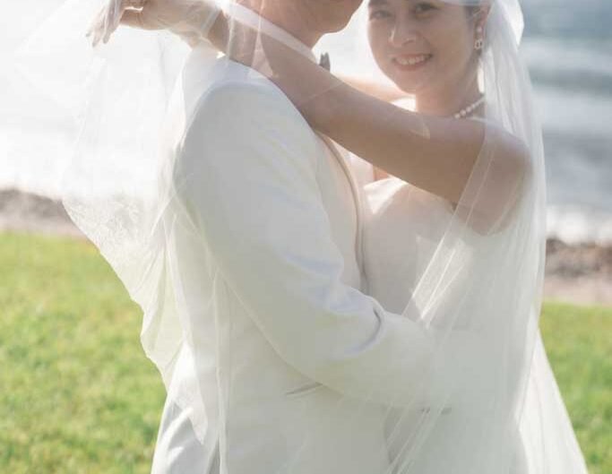 A close-up of a smiling bride and groom enveloped in a bridal veil with soft light filtering through, set against the backdrop of Lake Como.