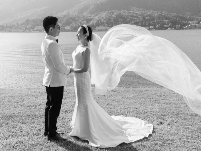 A black and white photograph of a bride and groom holding hands by Lake Como, with the bride's veil flowing in the wind and the mountains in the backdrop.