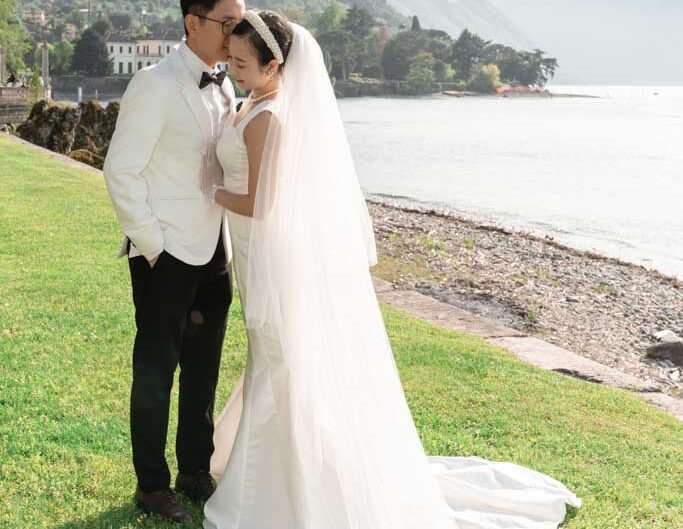 A bride and groom stand forehead to forehead, enveloped in the bride’s long veil, with the sunlit Lake Como and mountainous landscape in the background.