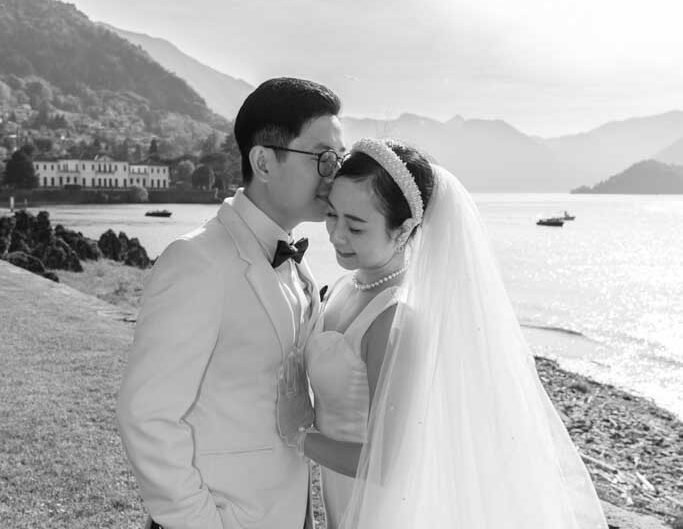 A black and white image of a bride and groom sharing a close, affectionate moment by the shore of Lake Como, with mountains in the backdrop.