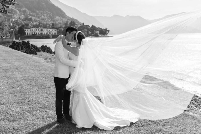 n a black and white photo, a bride and groom share a kiss by Lake Como, the bride’s veil caught in a gentle breeze, with the picturesque landscape and villa in the background.
