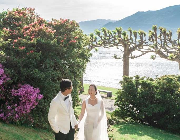 A bride in a white gown and a groom in a white tuxedo hold hands in a lush garden, with a sparkling lake and mountains under a bright sky in the background.