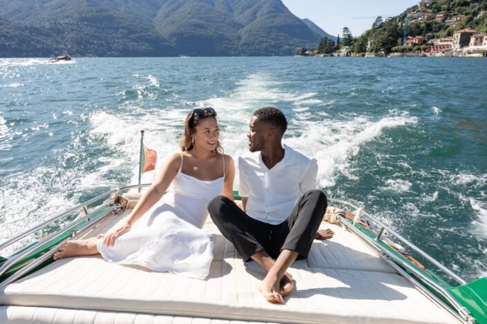 A man and a woman, both smiling and dressed elegantly in white, are sitting closely and looking at each other on a boat, with an Italian flag fluttering in the breeze and the waters of Lake Como splashing around them