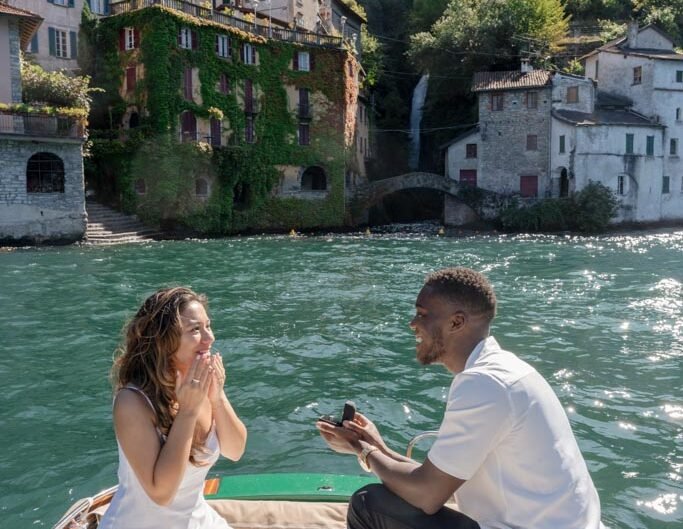 A woman reacts with joyful surprise as a man proposes to her on a boat on Lake Como, with historic buildings in the background