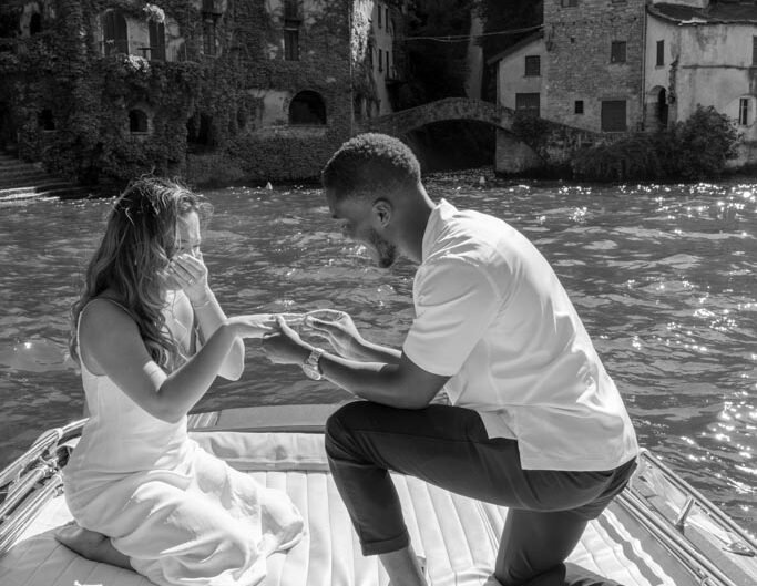 "In a black-and-white photo, a man kneels on one knee on a boat, offering a ring to a woman against the backdrop of historical lakeside buildings