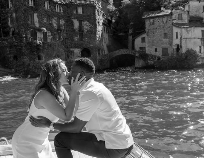 A black-and-white photo of a couple embracing on a boat on Lake Como after a proposal, with historic buildings in the background