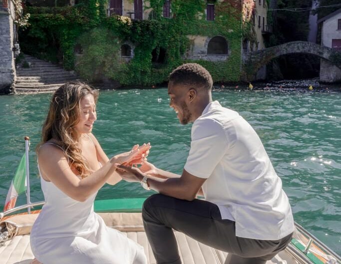 A man on one knee proposes to a woman on a boat on Lake Como, with ivy-clad buildings in the background