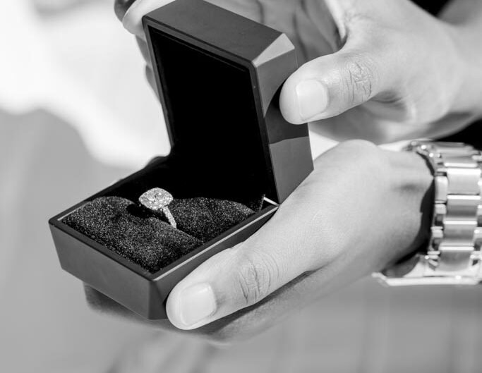 A close-up in black and white of a person's hands holding an open ring box with a sparkling engagement ring inside