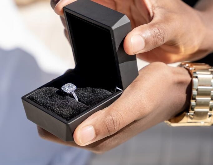 A close-up in black and white of a person's hands holding an open ring box with a sparkling engagement ring inside