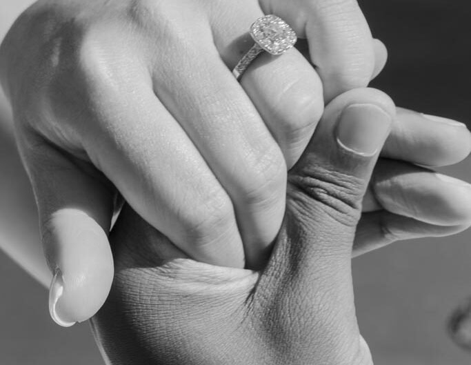 A black-and-white image showing two hands clasped together, with one hand showcasing a diamond engagement ring