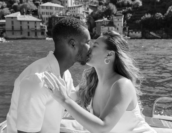 A black-and-white image of a couple sharing a kiss on a boat with the picturesque backdrop of Lake Como's hillside buildings