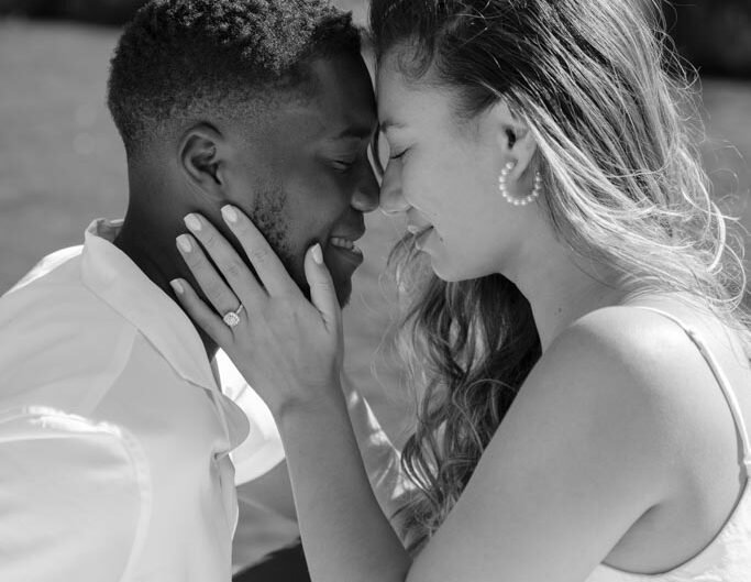 A black and white image of an intimate moment between a newly engaged couple, foreheads touching and smiling gently