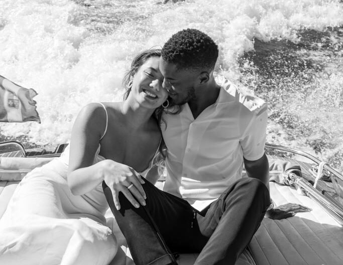 A black-and-white image of a smiling couple in an affectionate embrace on a boat, with water churning behind them, indicating a proposal has just taken place on Lake Como