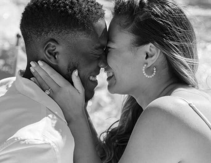A close-up in black and white of a couple intimately touching foreheads, smiling with their eyes closed