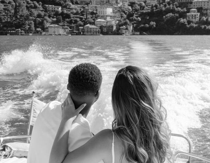 A black and white image of a couple seated closely together on a boat, looking out over the waves of Lake Como.