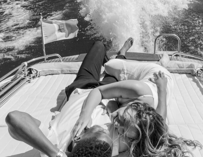 A black and white image of a couple lying on a boat, joyfully intertwined, with Lake Como’s waters churning behind them