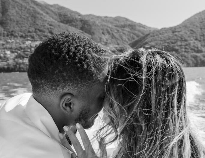 In a black and white photograph, a couple shares an intimate moment on a boat, with Lake Como's mountains in the backdrop