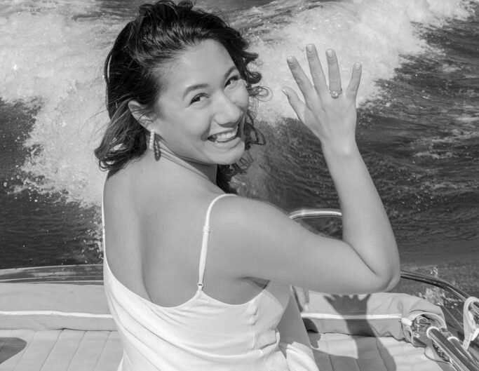A black and white image of a woman showing off her engagement ring, smiling broadly on a boat, with Lake Como in the background