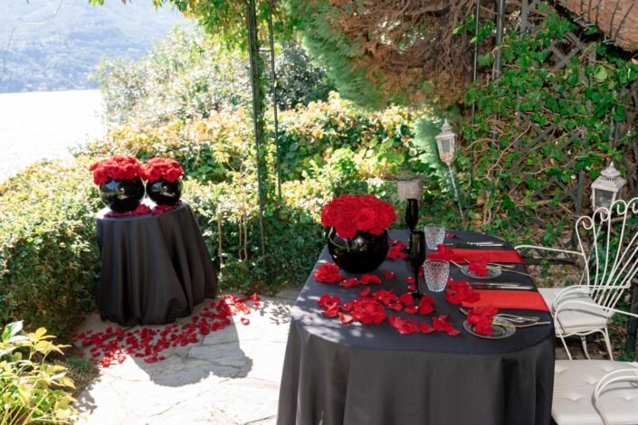 An elegant outdoor dining setup with red roses and petals scattered on a black tablecloth, overlooking Lake Como