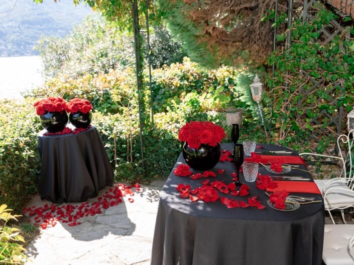 An elegant outdoor dining setup with red roses and petals scattered on a black tablecloth, overlooking Lake Como