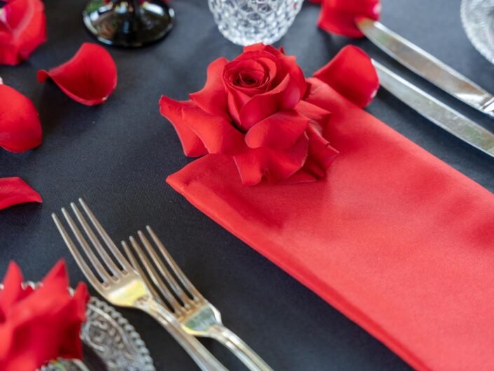 A meticulously set dining table with a single red rose on a napkin and rose petals scattered around