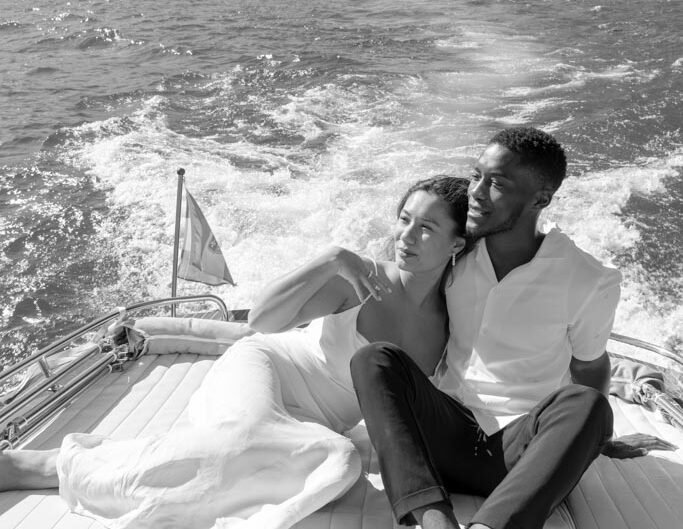 In a black-and-white photo, a couple shares a tender moment on a boat, the woman resting on the man's chest, with the backdrop of Lake Como’s mountains and village