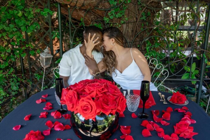 A couple lovingly touches foreheads, surrounded by a romantic proposal setting with red roses and elegant tableware.