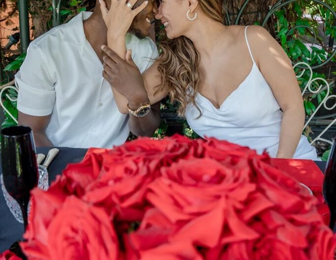 A newly engaged couple share a moment of affection, surrounded by a lavish display of red roses, during a proposal in Lake Como