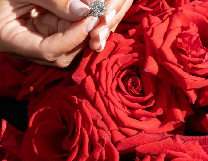 A close-up of a hand delicately holding an engagement ring against a backdrop of vibrant red roses