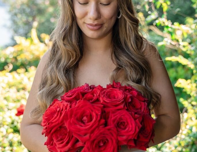 A woman in a white dress, eyes closed, holds a large bouquet of red roses, embodying a moment of blissful anticipation