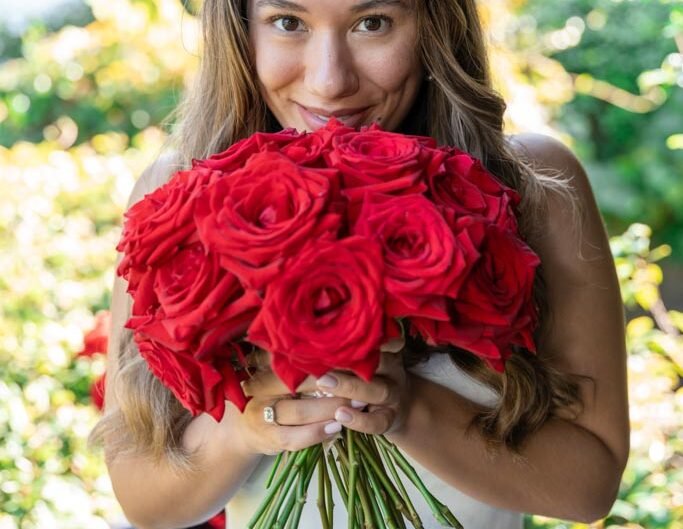 A smiling woman in a white dress peeks over a vibrant bouquet of red roses, her eyes gleaming with happiness