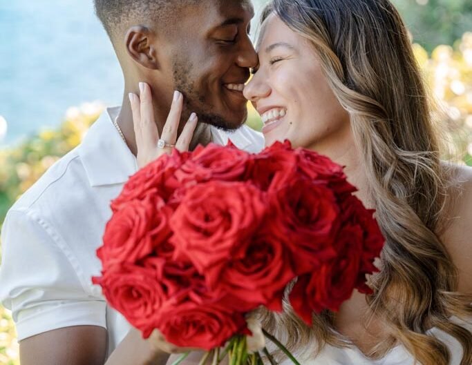 A couple in white clothing shares a close, joyful moment with a large bouquet of red roses partially obscuring their faces