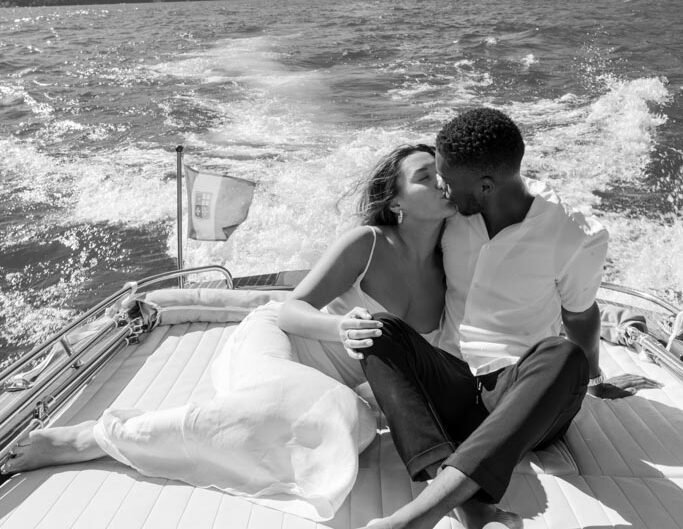 A black-and-white photo of a couple in a close embrace on a boat at Lake Como, with mountainous scenery in the background